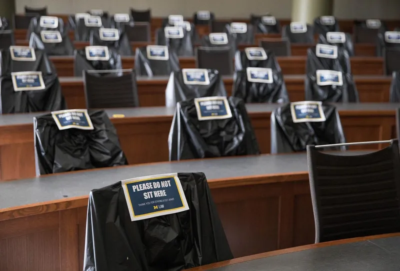 Chairs in a Law School lecture hall are covered in plastic with signs that read "Do Not Sit Here."