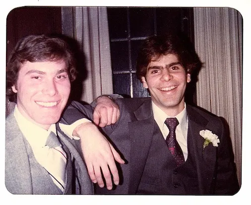 Two young men in suits smile with their arms around each other.