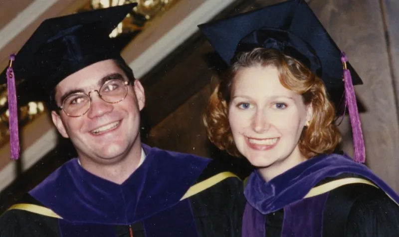 A bespectacled man and woman with shoulder-length hair smile in graduation gowns and caps. 