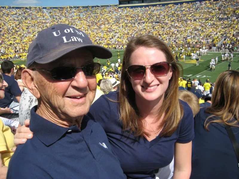 A woman in sunglasses puts her arm around an older man with a Michigan football game in progress behind them.