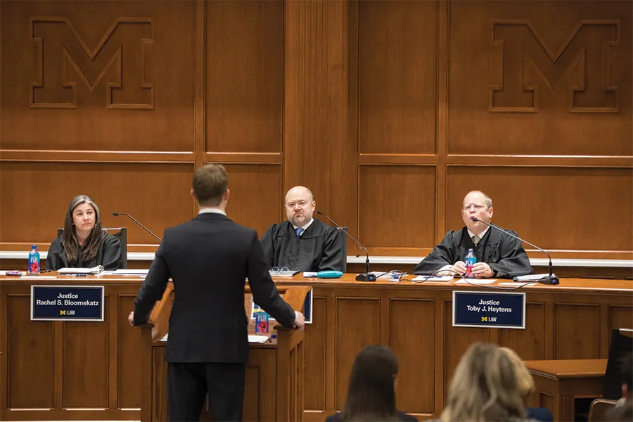 summer-2025-cover-gallery-color-014.jpg A man at a podium speaks to a panel of three judges.