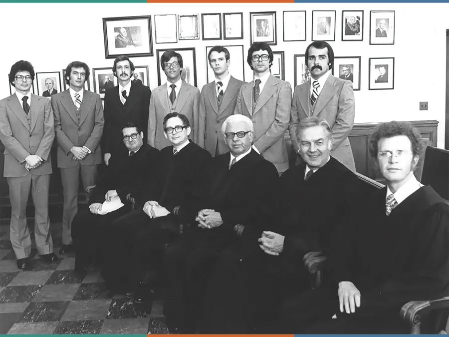 A row of formally dressed law students stand behind a row of seated judges in robes. 