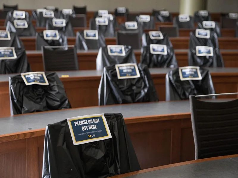 Chairs in a Law School lecture hall are covered in plastic with signs that read "Do Not Sit Here."