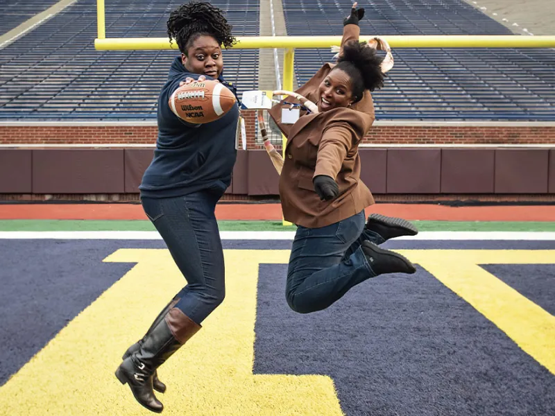 Two people one with the football, jumping up on the football field at the  UM Bar Alumni Reception at the Big House