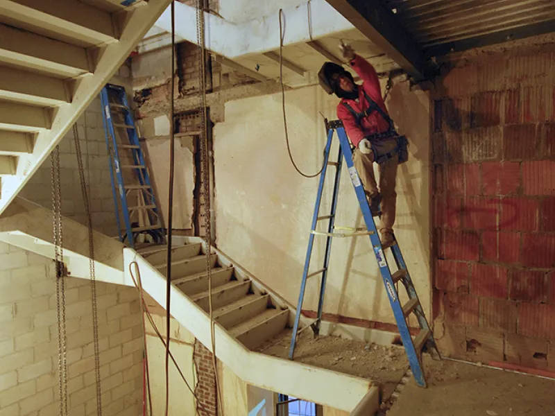 A construction worker on a ladder works on a ceiling while perched within a stairwell.