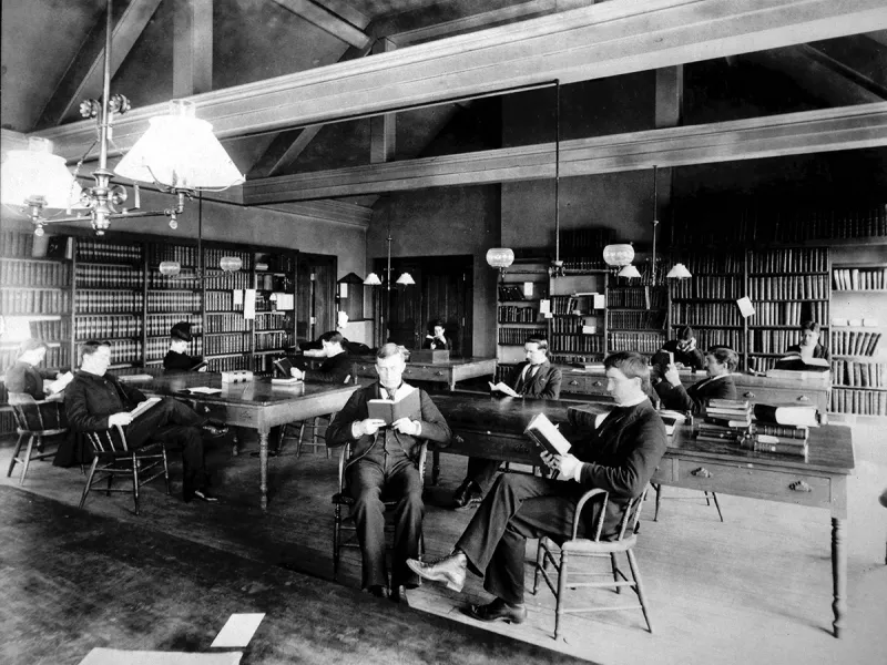 Men in suits sit at wooden tables surrounded by shelves of books in a high-ceilinged library.