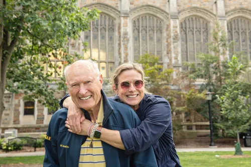 A woman in sunglasses wraps her arms around the shoulders of an older bald man in the sun of the Law Quad.