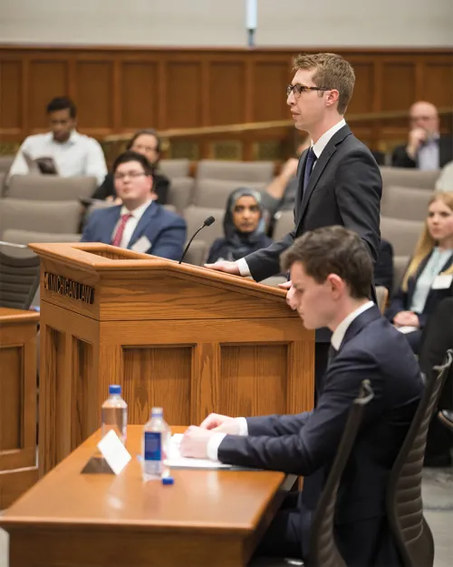 summer-2025-cover-feature-aside-001.jpg A man standing at a podium in an auditorium. Next to him, a man is seated at a desk reviewing documents.