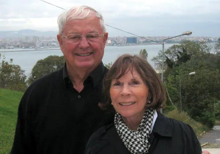 A man with white hair and his wife smile with a lake in the distance.