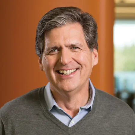 A man in a sweater and button-down smiles in front of a warm wood wall.