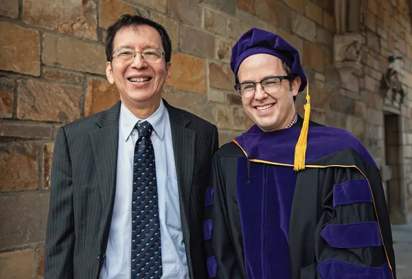 Two men, one in a suit and the other in a graduation gown, smile in front of a stone wall.