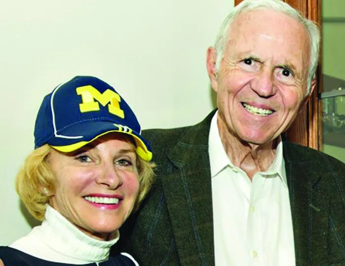 A woman with a Michigan hat and a turtleneck stands arm-in-arm with a man in a blazer.