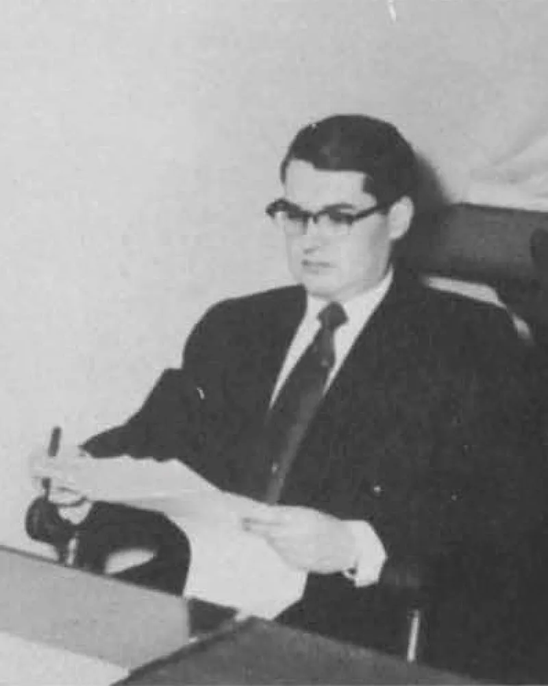 summer-2025-cover-feature-aside-009.jpg A formally dressed man in horn-rimmed glasses sits at a table with a notebook in his lap.