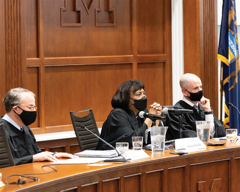 summer-2025-cover-feature-aside-014.jpg Three judges, wearing medical face masks, listen to someone who is speaking out of frame.