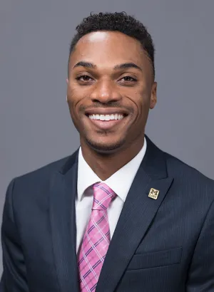 A person with close-cropped hair smiles for a professional photo, dressed in a suit and tie. 