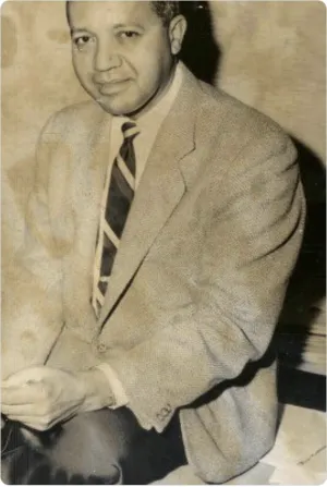 A man in a suit hunches against a wall with a smile on his face.