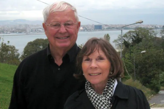 A man with white hair and his wife smile with a lake in the distance.