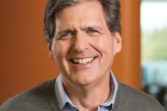 A man in a sweater and button-down smiles in front of a warm wood wall.