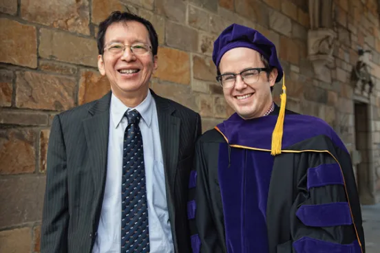 Two men, one in a suit and the other in a graduation gown, smile in front of a stone wall.