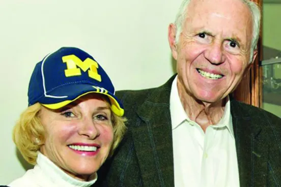 A woman with a Michigan hat and a turtleneck stands arm-in-arm with a man in a blazer.