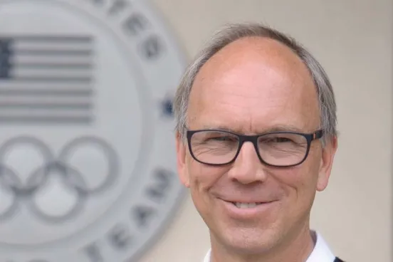 A man in glasses stands in front of a U.S. Olympics sign.