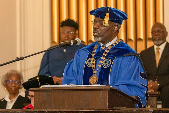 Clarence Armbrister in a blue robe and cap and gown speaking into a microphone