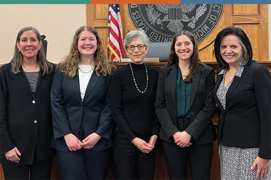 Two Michigan Law students with Michigan Law faculty and alumni A group of five in professional attire stand together in front of a wooden courtroom backdrop, with an American flag and court seal visible.