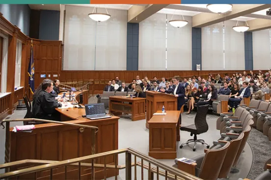 A mock trial being held at Michigan Law A courtroom scene shows a judge presiding over a case, with a lawyer presenting evidence to an attentive audience in a wooden-paneled space.
