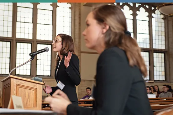 A student speaks at a podium in a spacious hall with large stained glass windows, while an audience listens attentively in the background.