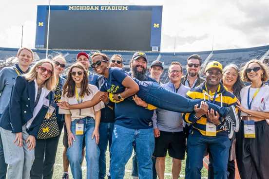 Ashley M. Washington (aloft), ’10, with classmates during a 2025 Reunion event at Michigan Stadium.