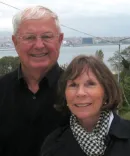 A man with white hair and his wife smile with a lake in the distance.