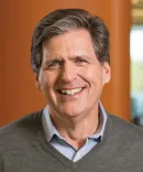 A man in a sweater and button-down smiles in front of a warm wood wall.