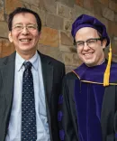 Two men, one in a suit and the other in a graduation gown, smile in front of a stone wall.