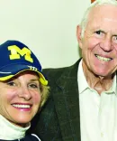 A woman with a Michigan hat and a turtleneck stands arm-in-arm with a man in a blazer.
