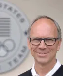 A man in glasses stands in front of a U.S. Olympics sign.