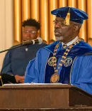 Clarence Armbrister in a blue robe and cap and gown speaking into a microphone