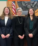 Two Michigan Law students with Michigan Law faculty and alumni A group of five in professional attire stand together in front of a wooden courtroom backdrop, with an American flag and court seal visible.
