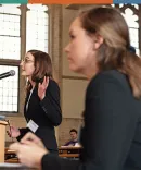 Michigan Law student during mock trial A student speaks at a podium in a spacious hall with large stained glass windows, while an audience listens attentively in the background.